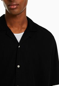 Close-up of a man wearing a black button-up shirt over a white undershirt, showing the neck, shoulders, and upper chest.