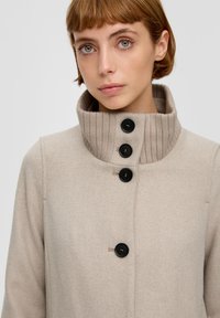 Beige jas met een geribbelde hoge kraag, voorzien van drie zwarte knopen en een subtiele visgraatstructuur. Gladde stofafwerking.