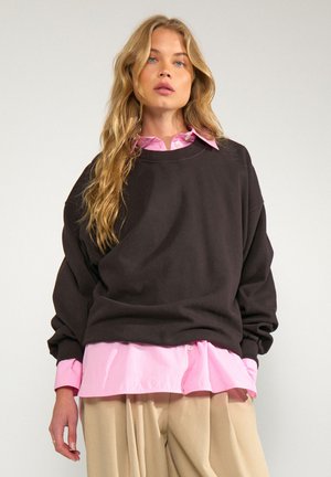 Donna bionda che indossa un maglione nero oversize sopra una camicia rosa e pantaloni beige, in piedi davanti a uno sfondo chiaro e semplice.