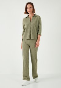 Chemise boutonnée en vert olive texturé avec des manches retroussées, associée à un pantalon assorti. Des baskets blanches complètent la tenue.