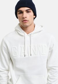 Sudadera blanca con el logo "GANT" en relieve, capucha con cordones y bolsillo frontal. Llevada con un gorro de punto azul oscuro. Textura suave y lisa.