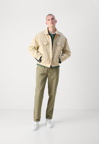 Beige Jeansjacke, grüner Pullover darunter, olivfarbene Hose und weiße Sneakers. Zwei Brusttaschen, Knopfverschlüsse an der Jacke, gerade Passform.