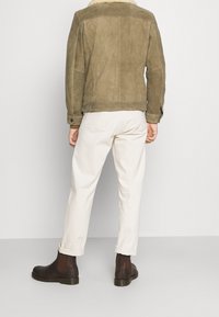 Hnědá semišová bunda s límcem z beránčí kožešiny, doplněná krémovými kalhotami a tmavě hnědými kotníkovými botami. Hladká textura a přiléhavý střih.