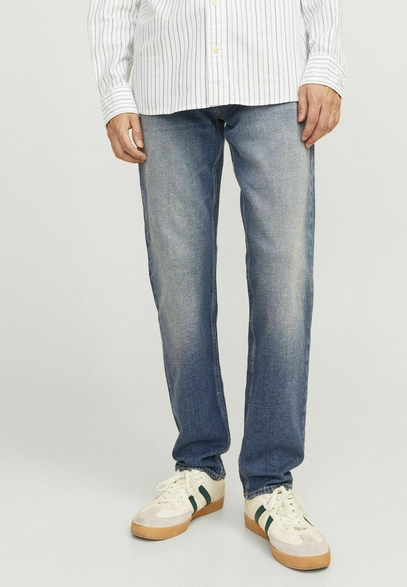 Jeans de mezclilla azul claro con un acabado desgastado, diseño de pierna recta y estilo clásico de cinco bolsillos, combinados con zapatillas blancas con rayas verdes.