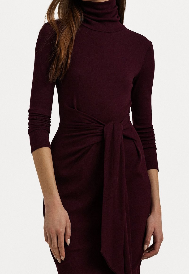 Femme portant une robe col roulé à manches longues couleur violet foncé avec un nœud lié à la taille sur un fond uni.