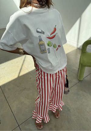 Persoană purtând un tricou alb oversized cu grafice de chili, lime, tequila și sare, asortat cu pantaloni evazați cu volane, în dungi verticale roșu și alb.