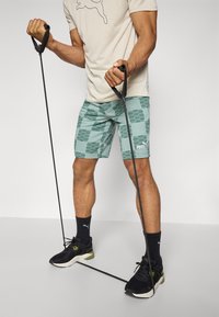 Calções de atletismo Puma em azul-turquesa com um padrão geométrico, combinados com uma t-shirt bege e ténis pretos. O utilizador segura fitas de resistência.
