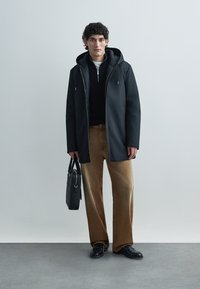 Crna jakna s kapuljačom i patentnim zatvaračem sprijeda, u kombinaciji s crnim džemperom i smeđim širokim hlačama od manšestru. Model drži crnu torbu.