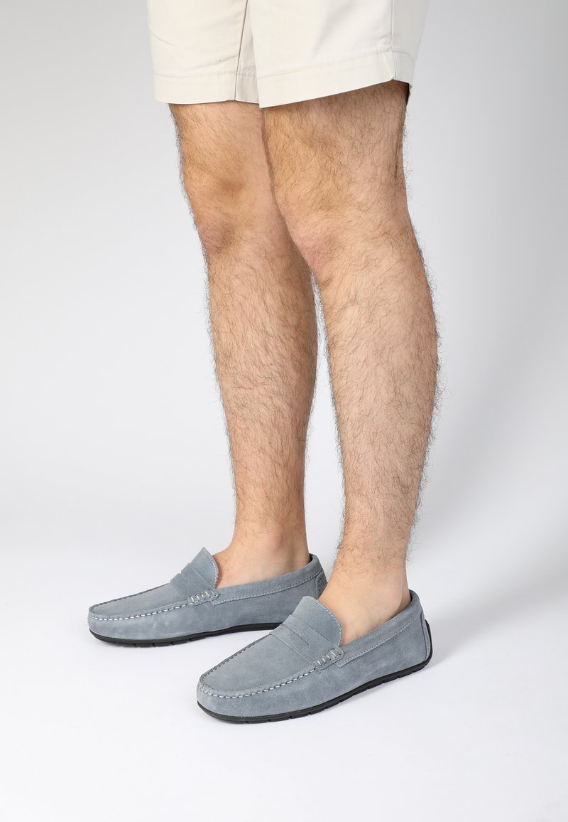 Graue Wildleder-Mokassins im Loafer-Stil mit einer flachen schwarzen Sohle, die über eine gestickte Akzentuierung und ein offenes Rückendesign verfügt.