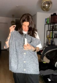 Persona con capelli scuri esamina un cardigan grigio con bottoni all'interno, circondata da scaffali, libri e abbigliamento in un ambiente soggiorno.