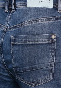 Mörkblå denimjeans med en bakficka som har en silverknapp och subtila sömnadsdetaljer. Etikett vid midjan i ljusblå.