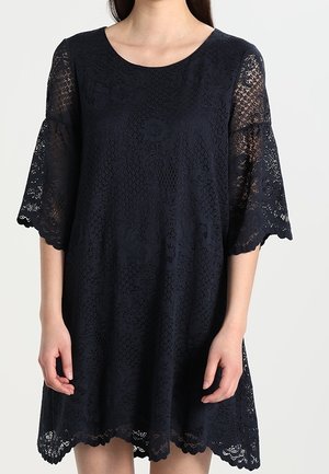 Kobieta ubrana w granatową koronkową sukienkę z rozszerzanymi rękawami do łokcia i falowanym brzegiem, stojąca na tle jednolitego tła.