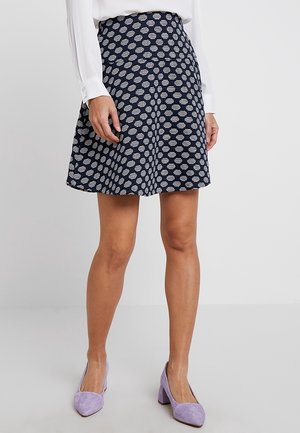 Vrouw die een marineblauwe rok met witte cirkelpatronen, een wit shirt en lavendel suède lage schoenen met blokhak draagt, staand op een lichte vloer.