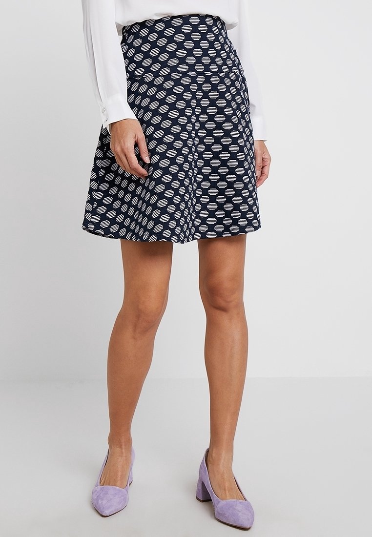 Femme portant une jupe bleu marine avec des motifs circulaires blancs, une blouse blanche, et des chaussures en daim lavande à petit talon carré, debout sur un sol clair.