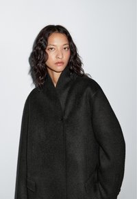 Czarny wełniany płaszcz z wysokim kołnierzem i zapięciem na jeden guzik. Charakteryzuje się luźnym krojem i minimalistycznym stylem.