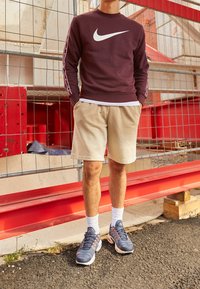 Maroon Nike sweatshirt with white logo, beige shorts, and navy athletic shoes. Sweatshirt has white and red accents on sleeves. White socks visible.