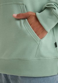 Sudadera verde clara con puños y dobladillo acanalados, que cuenta con un bolsillo frontal. Tela suave con una textura lisa y una etiqueta de marca minimal visible.