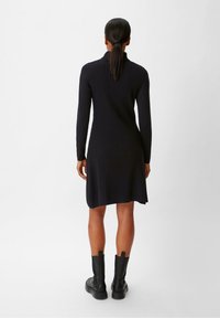 Robe noire en maille côtelée avec manches longues, col montant et ourlet évasé, portée avec des bottes noires jusqu'aux genoux. Texture lisse et coupe ajustée.