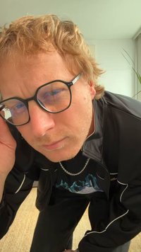 Μαύρο αθλητικό μπουφάν με λευκές λεπτομέρειες πάνω από μαύρο μπλουζάκι. Πάρα πολύ μαύρα γυαλιά με ασημένια λεπτομέρεια. Κοντά, ατημέλητα, ανοιχτό καφέ μαλλιά.