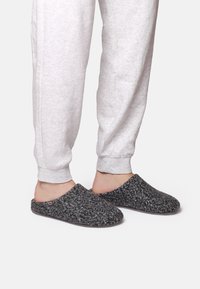 Grå texturerade tofflor med ett mjukt, plyschigt material och öppen baksida, bärs med ljusgrå sweatpants.