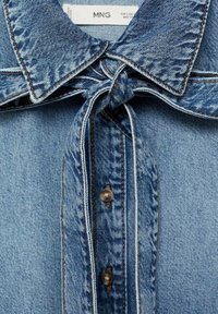 Camicia in denim azzurro chiaro con colletto annodato, apertura con bottoni e dettagli di cucitura a contrasto, con una texture liscia e un design classico.