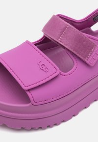 Lilla UGG suss, millel on reguleeritavad rihmad, pehme tallaga ja tekstuuriga jalapadi mugavuse ja haardumise tagamiseks.