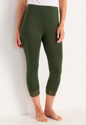 Kobieta w oliwkowo-zielonych, krótkich legginsach z koronkową lamówką, stojąca boso przy białej ścianie.