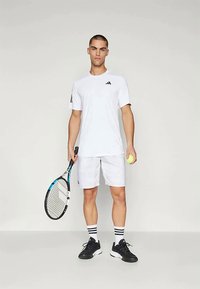 Camisa atlética de manga corta blanca con detalles negros, combinada con pantalones cortos blancos y zapatillas negras. Incluye una raqueta de tenis blanca y una pelota amarilla.