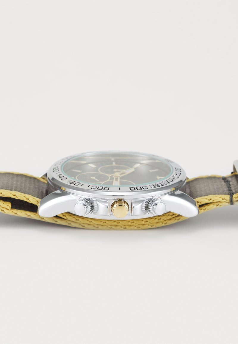 Montre en argent avec un cadran rond, un cadran vert, trois sous-cadrans, une couronne dorée et un bracelet en cuir texturé jaune avec des accents gris.