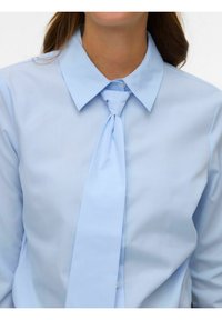 Camisa azul claro con cuello y corbata. La tela es lisa y la corbata tiene un nudo. La camisa tiene botones decorativos debajo del cuello.