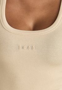 Beige ribbad linne med vid halsringning, med ett präglat "BOSS"-logotyp i mitten. Tyget ser mjukt och åtsittande ut.