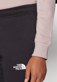 Čierne teplákové nohavice s elastickým pásom, na ľavom stehne s bielym logom The North Face. Rúžová tričko s dlhým rukávom je čiastočne viditeľné.