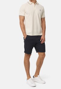 Mies, joka on pukeutunut vaaleanbeigeeseen poolopaitaan, tummiin lyhyisiin shortseihin, jotka on käännetty ylös, ja valkoisiin tennareihin, seisoo kädet taskuissa valkoista taustaa vasten.