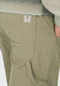 Olivengrønne fløjlbukser med ribbet tekstur, sidelommer og en label med et geometrisk design syet på baglommen.