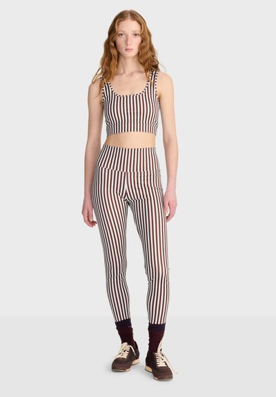 Jeune femme debout portant un crop top rayé vertical marron et blanc et des leggings assortis avec des chaussettes foncées et des baskets marron.