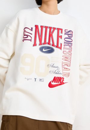 Personne portant un sweat-shirt blanc avec des logos Nike rouges et bleus, texte "1972," "90," et "Sportswear" sur le devant.