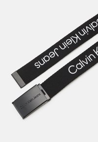 Zwarte stoffen riem met de tekst "Calvin Klein Jeans" in het wit, met een rechthoekige zwarte metalen gesp. De tekst is over de riem gedrukt.