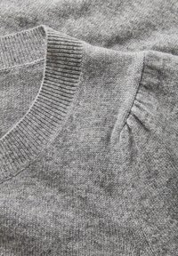 Gros plan sur un tissu de pull en tricot gris doux, montrant un col en V côtelé et un détail froncé à la couture de l'épaule.