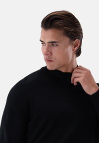 Schwarzer Rollkragenpullover, glatte Textur, taillierte Passform und gerippter Kragen. Das Model wirkt elegant und berührt mit einer Hand den Halsauschnitt.
