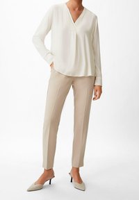 Blouse ivoire à manches longues avec un col en V et une poche, associée à un pantalon beige fuselé et des talons pointus de couleur claire.
