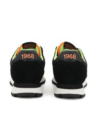 Schwarze Sneaker mit neongrünen und orangefarbenen Akzenten, weißen Sohlen, von hinten betrachtet, mit "1968" in Orange auf jeder Ferse gedruckt.