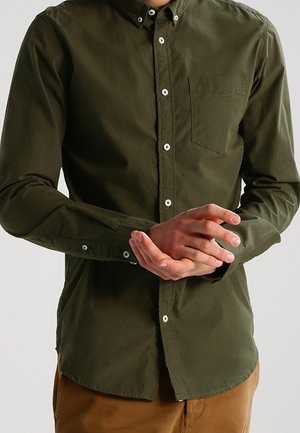 Person trägt ein olivgrünes Hemd mit Brusttasche und braune Hose, hält die Hände auf Taillenhöhe zusammen.