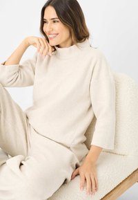 Cremefarvet fleece-top med høj krave og trekvartÆrmer, parret med matchende bløde bukser, på en tekstureret lys stol.
