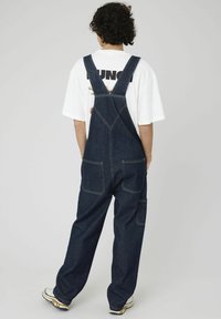 Salopette en denim bleu foncé avec des bretelles réglables, de grandes poches avant et arrière, et une coupe décontractée. Portée par-dessus un t-shirt graphique blanc.
