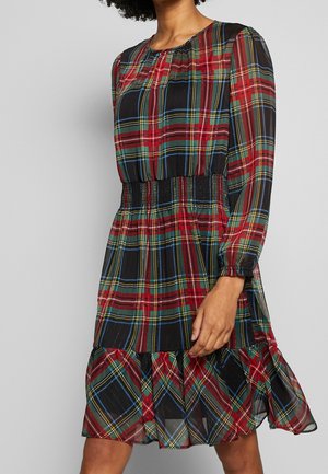 Vrouw draagt een jurk met lange mouwen tot op de knieën, met een ruitpatroon in rood, groen, blauw en geel en een getailleerde taille.