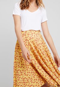 Keltainen kukkakuosinen hame, jossa on punaisia ja valkoisia kuvioita, yhdistettynä yksinkertaiseen valkoiseen V-kaulaiseen t-paitaan. Hame on liehuva ja hieman levenevä.