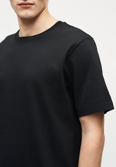 Νεαρό άτομο που φοράει μια απλή μαύρη κοντομάνικη μπλούζα με στρογγυλή λαιμόκοψη, μπροστά σε ουδέτερο φόντο, με την εικόνα περικομμένη κάτω από τη μύτη.