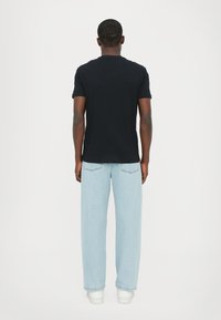 Svart t-skjorte med korte ermer kombinert med lyseblå vide jeans og hvite sko, fanget fra baksiden mot en ensfarget bakgrunn.