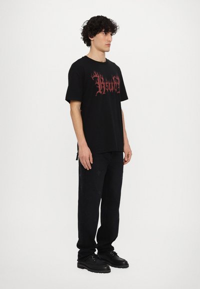 T-shirt noir avec un motif graphique en flamme rouge, associé à un pantalon noir avec des motifs croisés subtils et des chaussures noires. Vêtements décontractés.