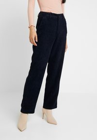 Mörkblå kordflärdsbyxor med vertikala ränder, elastisk midja, framfickor och avslappnad passform, tillsammans med beige spetsiga klackar.